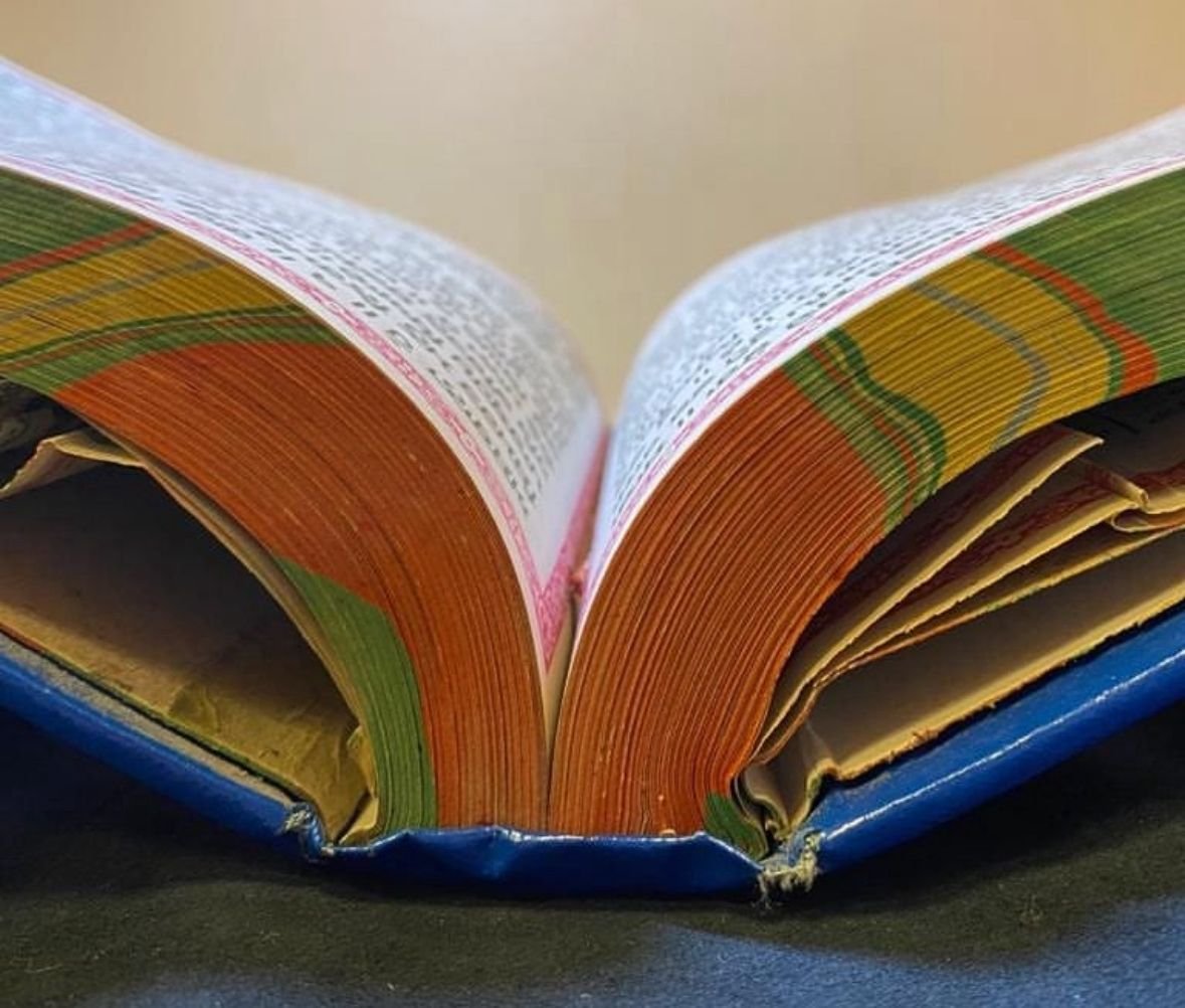 Close-up photograph of a Pothi Sahib showing pleated and torn endleaves at the spine gutter, with a blue cover and colourful striped pages
